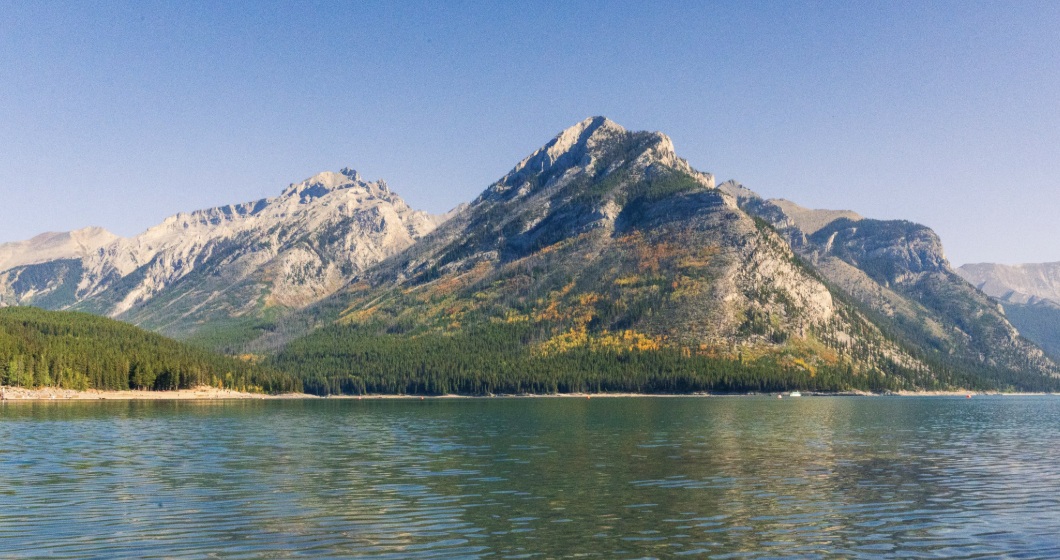 Sunlit mountains above a clear lake, with ripples and stones visible in the water